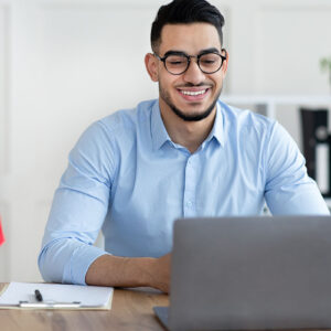 A smiling man wearing glasses sits at a desk with a laptop, engaged in online learning.