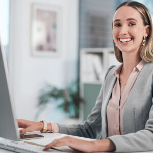 A woman with a smile sitting at a desk, using a computer.