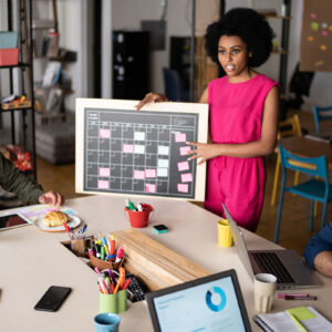 A woman holding a board with some sticky notes on it at a table with her coworkers, doing project management.