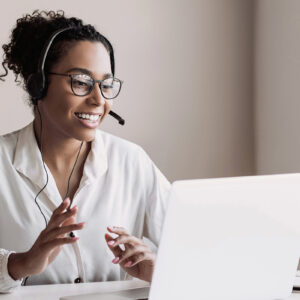 A woman with headset and glasses sitting at desk with laptop, working in a modern office setting.