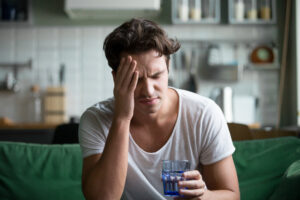 Young man suffering from headache at home, with his hand on his head and holding a glass of water
